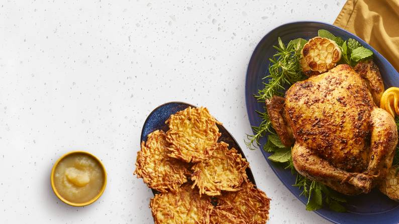 A roasted chicken on a platter with herbs, citrus, and garlic, a plate of potato pancakes, and a bowl of applesauce on a white granite countertop.
